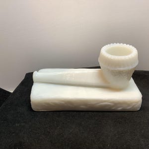 May include: A white milk glass pipe-shaped object resting on a rectangular base. The bowl of the pipe features a beaded rim and embossed decorative patterns. The stem is smooth and cylindrical, while the base displays a textured leaf-like motif.