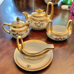Peut inclure: Service à thé vintage avec théière, crémier, sucrier et saucière. L'ensemble présente une base blanche avec des bordures dorées et des motifs floraux. La théière a une anse arquée et un bouton décoratif.