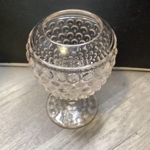 May include: A clear, glass footed bowl with a textured, hobnail pattern. The bowl has a round shape with a wide opening and sits on a short, rounded base. The hobnail design covers the entire bowl, creating a decorative effect.