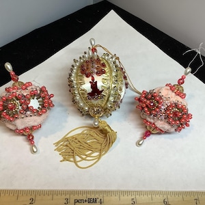 May include: Three decorative ornaments featuring pink and white fabric bases adorned with intricate red and gold beadwork. The central ornament includes a gold tassel and a candle motif. A ruler shows the set spans approximately 8 inches in width.