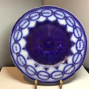 May include: A white and blue decorative plate with a scalloped edge. The plate features a central design with radiating lines and a monogram, surrounded by a border with the names of U.S. states. The plate is displayed on a gold stand.