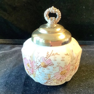 Puede incluir: Un tarro de cerámica blanca con un diseño floral y una tapa dorada con un asa plateada. El tarro tiene un borde festoneado y está decorado con flores rosas y amarillas.