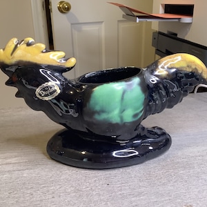 May include: A black and green ceramic rooster planter with a yellow glaze. The planter has a label that reads "Dryden".