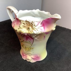 Pode incluir: Um cremeira de porcelana vintage com uma alça, apresentando um design floral em tons de rosa, amarelo e dourado. A cremeira tem uma borda com babados e uma aparência delicada pintada à mão, sugerindo um item antigo ou de colecionador.