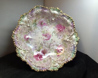 May include: A decorative, antique porcelain bowl with a ruffled edge. The bowl features a floral design with pink roses and green leaves on a white background. The rim is trimmed with gold.