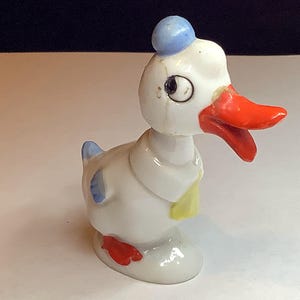 Puede incluir: Figura de pato de cerámica vintage, principalmente blanca con un pico naranja brillante y patas rojas. El pato lleva un sombrero azul y tiene detalles azules en las alas. Una pajarita amarilla añade un toque de color.