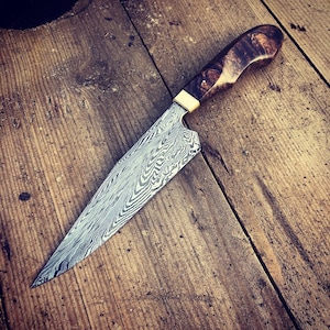 May include: A close-up of a kitchen knife with a wooden handle and a patterned blade. The blade is made of Damascus steel and has a unique, wavy pattern.