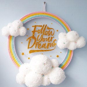 May include: A rainbow yarn wreath with the words "Follow Your Dreams" in gold glitter lettering. The wreath is decorated with three white fluffy pom-pom clouds.