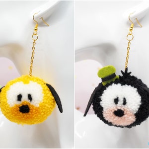 May include: A pair of dangle earrings featuring pom-pom characters. One earring is yellow with black ears and a black nose. The other earring is black with white eyes, a pink nose, and a green hat. Both earrings have gold chains.
