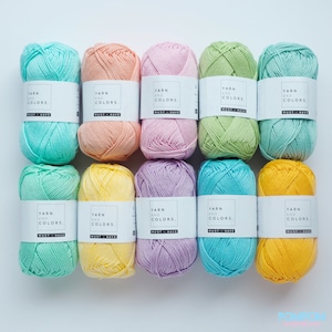 Könnte beinhalten: Zehn Knäuel Wolle in verschiedenen Pastellfarben, darunter hellblau, Pfirsich, Rosa, Grün und Gelb. Jedes Knäuel hat ein weißes Etikett mit der Aufschrift "Yarn and Colors. Must-Have."