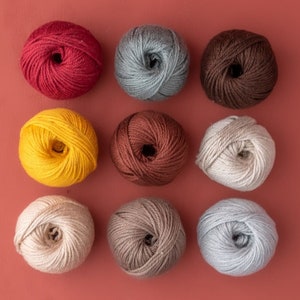 May include: Nine balls of yarn in various colours, including red, yellow, brown, grey, and beige. The yarn is arranged in three rows of three balls each.