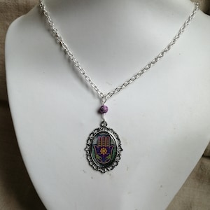 Può includere: Una collana a catena d'argento con un ciondolo a forma di mano di Hamsa viola e verde. Il ciondolo è in una cornice ovale con un bordo decorativo.
