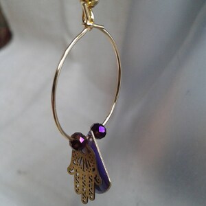 Può includere: Orecchini a cerchio dorati con un ciondolo hamsa viola e perline viola. Il ciondolo hamsa è una mano con dettagli intricati.