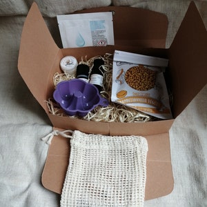 May include: A brown cardboard box filled with a variety of items, including a purple silicone mold, two small bottles, a white mesh bag, a small container of white cream, and a bag of yellow beads. The box is filled with shredded paper. The bag of beads has a label that reads "Emulsifiant Conditionneur Vegetal"