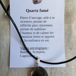 Peut inclure: Collier avec pendentif en quartz fumé sur un cordon noir. Le pendentif est de forme ovale gris foncé. La carte indique "Quartz fumé" et décrit les propriétés de la pierre. La carte liste également les signes astrologiques associés à la pierre : Balance, Scorpion, Capricorne.