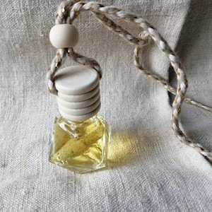 Op de afbeelding: Een kleine, heldere glazen fles gevuld met gele vloeistof, afgedekt met een houten dop en een gevlochten touw. Het touw is wit en goudkleurig. De fles is waarschijnlijk een etherische olie diffuser of luchtverfrisser.