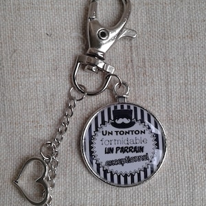 Może przedstawiać: Srebrny brelok do kluczy z okrągłym czarno-białym paskowanym brelokiem, na którym napisano "Un Tonton formidable Un Parrain exceptionnel". Brelok ma czarno-biały paskowany wzór z małym czarnym wąsem i kapeluszem cylindrycznym. Brelok ma również srebrny brelok w kształcie serca i gwiazdy.