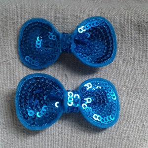 Op de afbeelding: Twee blauwe haarclips met een strik van pailletten. De strikken zijn gemaakt van blauwe pailletten en zijn bevestigd aan een clip.