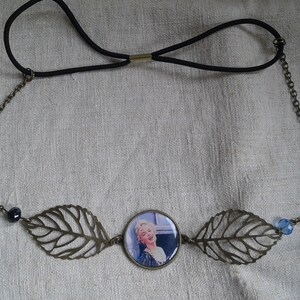 Op de afbeelding: Een bruin metalen hoofdband met een zwart elastiek. Het hoofdband heeft twee blaadjesvormige bedels aan weerszijden van een ronde bedel met een afbeelding van een vrouw. De bedels zijn met een ketting verbonden.