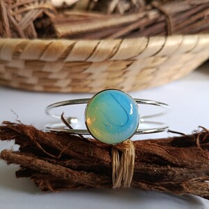 Peut inclure: Un bracelet argenté orné d'un grand cabochon rond bleu et blanc. Le bracelet est présenté sur un fagot de brindilles brunes, avec un panier tressé en arrière-plan. Le cabochon présente un aspect marbré.
