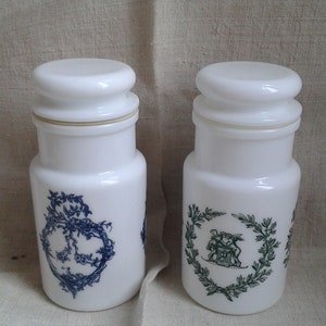 Peut inclure: Deux pots à pharmacie blancs avec couvercles assortis. L'un présente un motif floral bleu, l'autre une couronne verte avec une scène classique. Les pots sont cylindriques et reposent sur un tissu neutre.