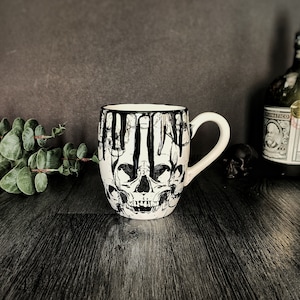 May include: White ceramic mug with a black and white skull design. The mug has black dripping paint around the rim and down the sides.