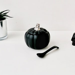 May include: Black ceramic pumpkin shaped sugar bowl with a silver lid. The bowl is sitting on a white surface next to a black ceramic skull and a silver spoon.