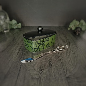 Puede incluir: Un recipiente para mantequilla de cerámica verde y negro con un patrón de serpiente y una tapa negra. Un cuchillo de mantequilla de esqueleto plateado está al lado del plato.