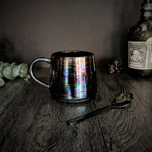 May include: A black iridescent ceramic mug with a handle. The mug is sitting on a dark wooden surface with a black metal spoon with a skull design.