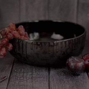 May include: A black ceramic bowl with a dripping skull design, filled with red grapes and surrounded by plums on a dark wood surface.