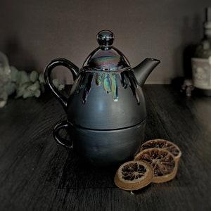 May include: A black ceramic teapot with an iridescent glaze and a matching teacup. The teapot has a drip-style design and a lid with a knob. The teacup has a handle.
