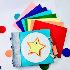 May include: A blue felt book with a yellow star and a white circle in the centre. The book is open to reveal a colourful felt rainbow with different colours of felt.