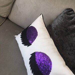 May include: White rectangular pillow with a black and purple sequined eye design. The pillow has a black border and is resting on a gray sofa. The purple sequins create a sparkling effect.