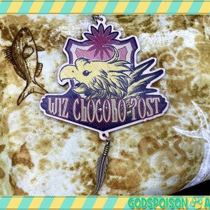 Könnte beinhalten: Ein Holzanhänger mit einem rosa-gelben Vogeldesign, dem Text "WIZ CHOсOBO POST" und einem Federschmuck. Der Anhänger befindet sich auf einem braunen und weißen Stoffhintergrund.