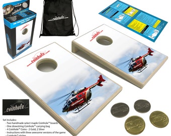 May include: A set of two wooden coin toss game boards featuring a red and white helicopter graphic. The boards are white with a brown trim and a circular hole in the centre. The set includes a drawstring bag, four coins, and instructions. The text on the boards reads "www.coinhole.com" and "NIGHTINGALE".