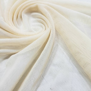 May include: Close-up of a cream-colored mesh fabric with a textured, woven pattern. The fabric is folded and draped, creating a soft, flowing look.