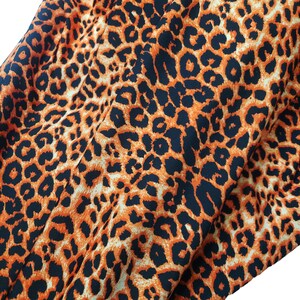 Peut inclure: Gros plan sur un tissu à motif léopard. Le tissu présente un motif répété de taches noires sur un fond beige et orange. Le tissu est plié, montrant la texture et le drapé du matériau.