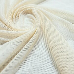 Pu&ograve; includere: Primo piano di un tessuto a rete color crema con un motivo testurizzato a griglia. Il tessuto &egrave; piegato e drappeggiato, creando curve e pieghe morbide.