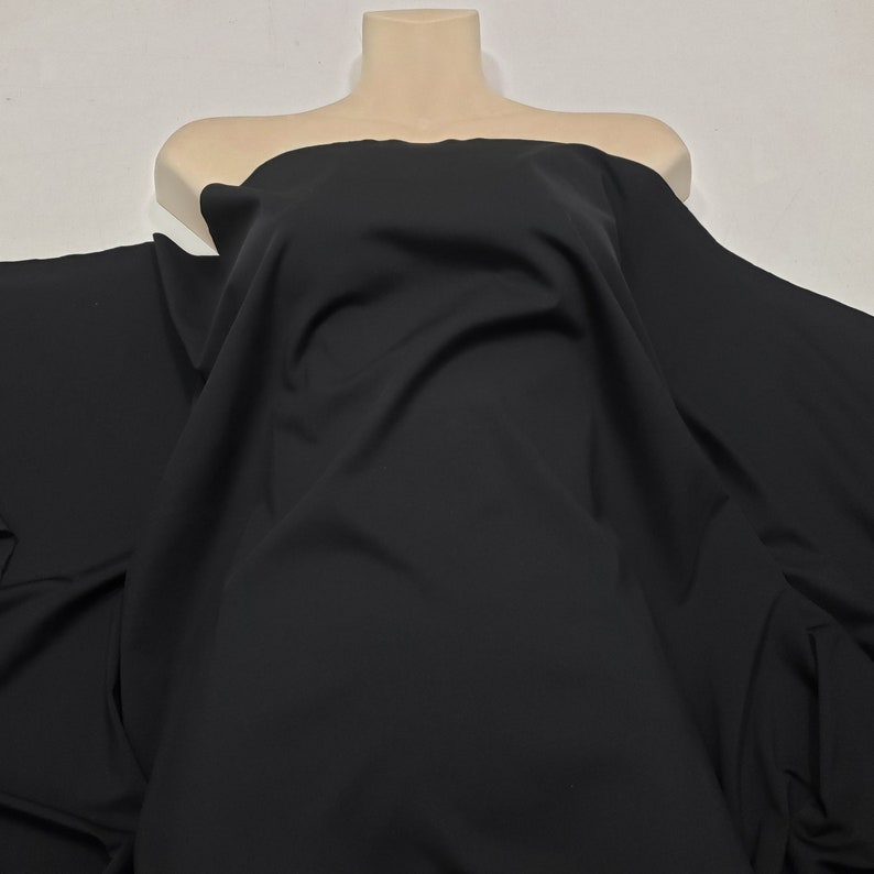 May include: A large piece of black fabric draped over a mannequin torso. The fabric is smooth and appears to be a solid color, with soft folds and shadows. The material is likely used for clothing or other textile projects.