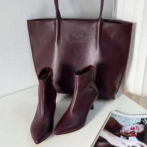 May include: A burgundy tote bag and matching ankle boots with pointed toes and stiletto heels. The bag has a rectangular shape with a small rectangular logo. A magazine is visible in the bottom right corner.