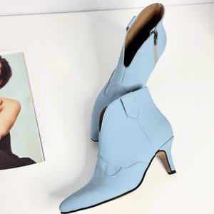 May include: A pair of light blue ankle boots with a pointed toe and a small heel. The boots have a western-inspired design with decorative stitching and a side zipper. The heel measures approximately 2.5 inches.