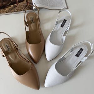 May include: Four pairs of women's slingback flats in neutral colors. Two pairs are a light tan, and two are white. Each shoe has a pointed toe and a thin strap around the ankle. The brand name "Klavels" is printed on a black label.