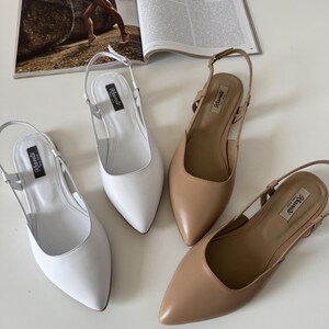 May include: Four pairs of women's slingback flats in white and tan leather. The shoes have pointed toes and adjustable ankle straps. The shoes are arranged on a white surface, with a magazine in the background.