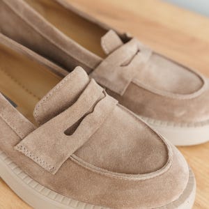 May include: A pair of light brown suede loafers with a white rubber sole. The loafers have a classic penny loafer design with a strap across the top.