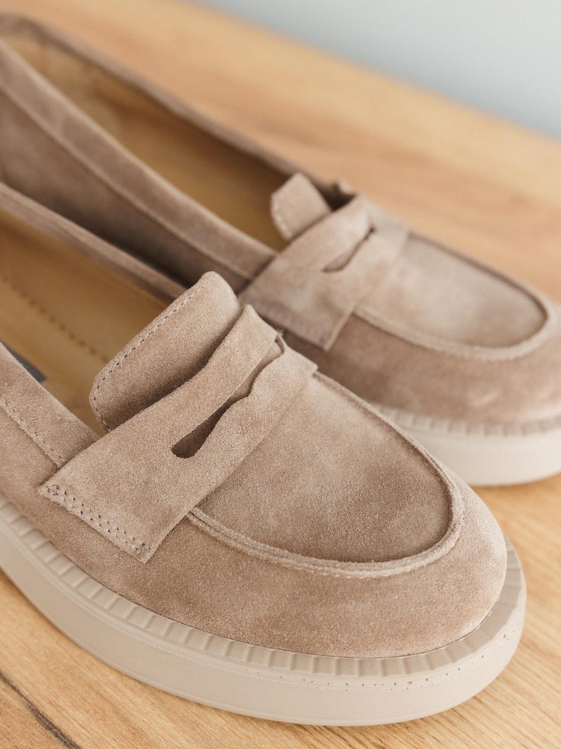 May include: A pair of light brown suede loafers with a white rubber sole. The loafers have a classic penny loafer design with a strap across the top.
