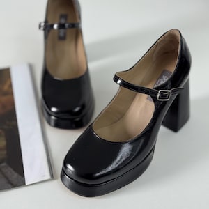May include: A pair of black patent leather platform shoes with a chunky heel and a buckle strap. The shoes have a square toe and a closed back.