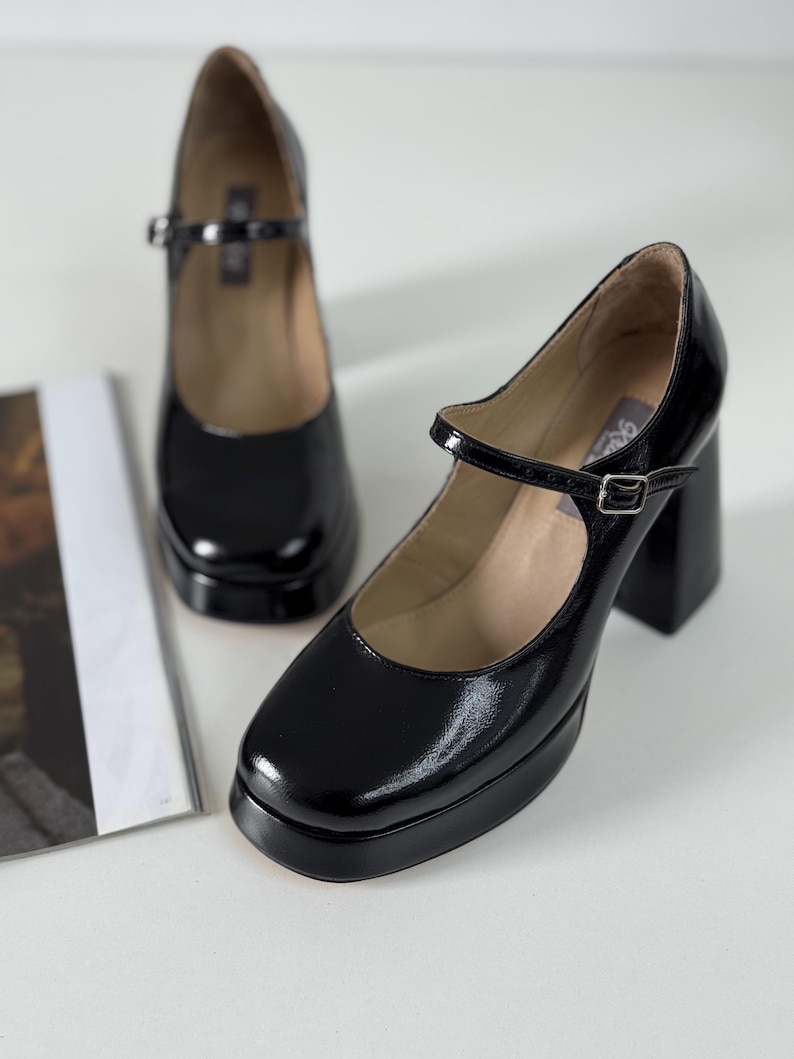 May include: A pair of black patent leather platform shoes with a chunky heel and a buckle strap. The shoes have a square toe and a closed back.