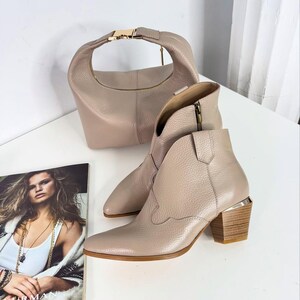 May include: A pair of beige leather ankle boots with a pointed toe and a stacked wooden heel. A matching beige handbag with a gold-tone clasp and zipper is also present. A magazine with a woman on the cover is in the foreground.