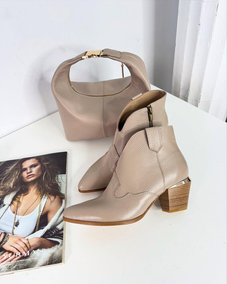 May include: A pair of beige leather ankle boots with a pointed toe and a stacked wooden heel. A matching beige handbag with a gold-tone clasp and zipper is also present. A magazine with a woman on the cover is in the foreground.