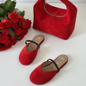 K&ouml;nnte beinhalten: Eine rote Wildlederhandtasche mit goldfarbener Schnalle und Rei&szlig;verschluss, kombiniert mit passenden roten Wildleder-Slippern mit schwarzen Riemen. Ein Strau&szlig; roter Rosen ist ebenfalls zu sehen.